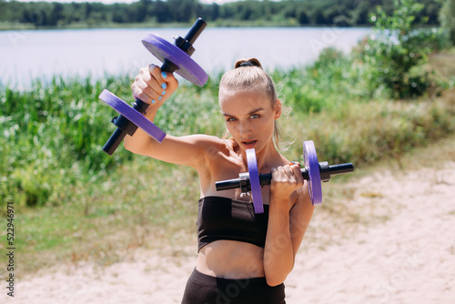 A tanned girl with a slim figure and blond hair, in black shorts and a tank top, is engaged in fitness with gartels on the beach, boxing, against the backdrop of a picturesque river in the summer. photo