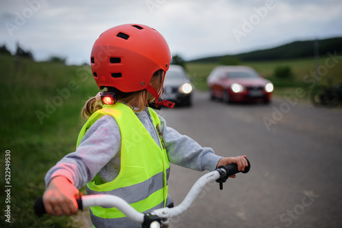Rear view of little girl in reflective vest riding bike on road with cars behind her, road safety education concept.