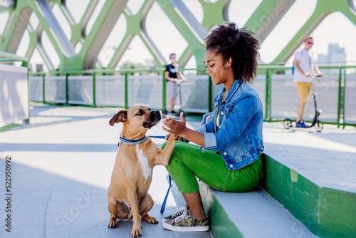 Fototapeta Naklejka Na Ścianę i Meble -  Multiracial girl sitting and resting with her dog outside in the bridge, training him, spending leisure time together. Concept of relationship between dog and teenager, everyday life with pet.