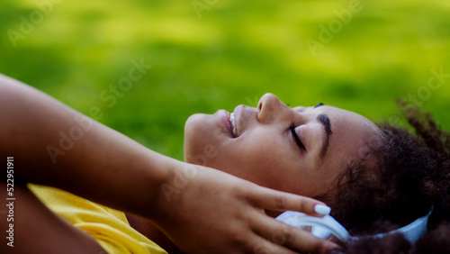 Multiracial girl lying down in grass and enjoying music in headphones  side view.