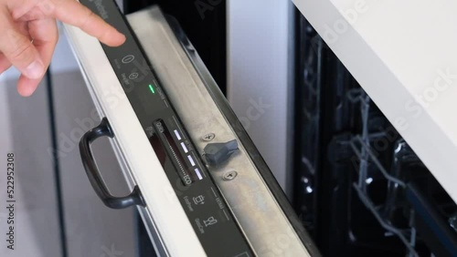 A finger is turning on a built-in dishwasher and selecting eco mode, closing the door.A woman hand is pressing the start button of built-in dishwasher control panel.Modern white kitchen. Close-up