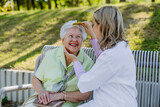Caregiver helping senior woman to comb hair and make hairstyle when sitting on bench in park in summer.