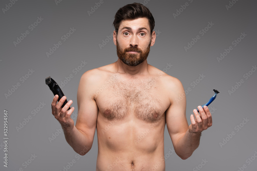 confused and shirtless man with beard holding safety and electric razors isolated on grey. confused and shirtless man with beard holding safety and electric razors isolated on grey.