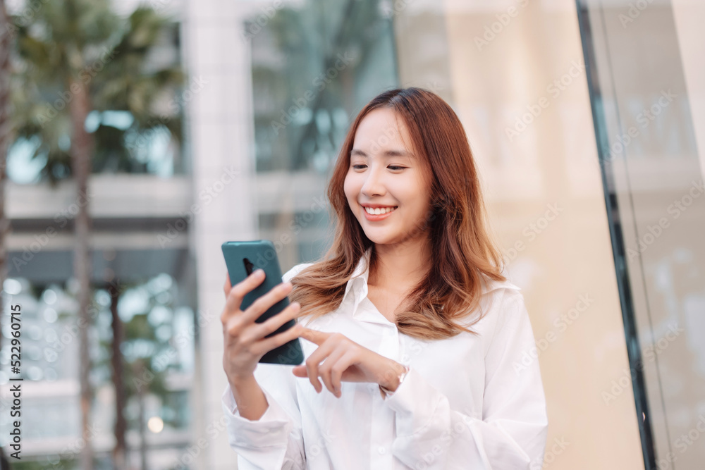 Fototapeta premium Beautiful asian female businesswoman use smartphone, Walk enjoy smiling while doing commuting in the modern city near office building outside.
