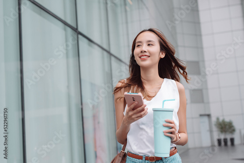 Smiling asian businesswoman use smartphone and hold reusable eco-friendly ecological cup while commuting in city in the morning