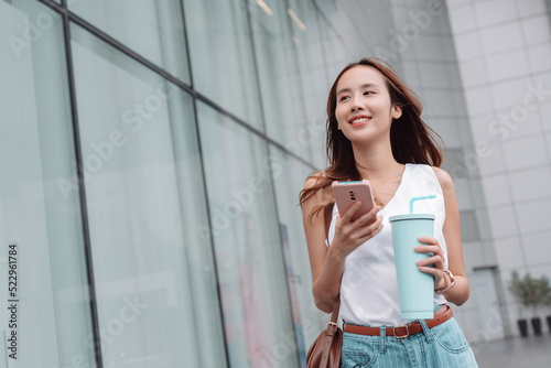Smiling asian businesswoman use smartphone and hold reusable eco-friendly ecological cup while commuting in city in the morning