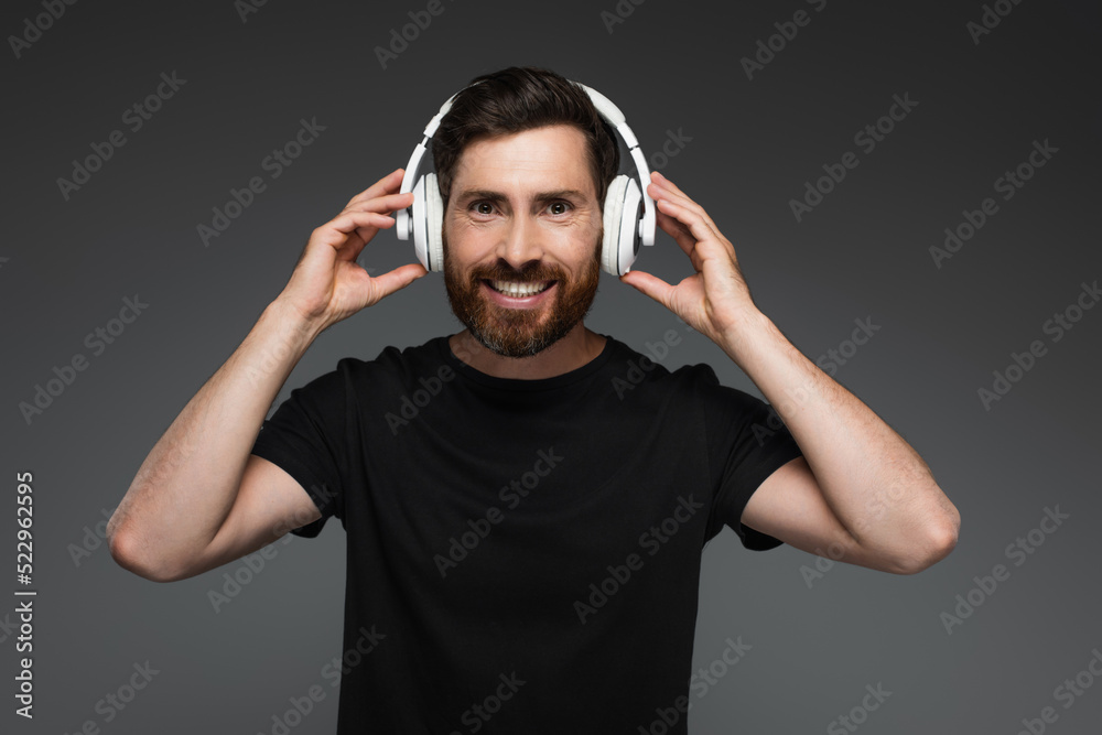 Naklejka premium cheerful and bearded man in wireless headphones listening music isolated on grey.