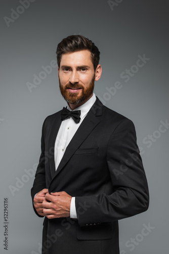 happy and successful man with beard standing in elegant suit with bow tie isolated on grey.