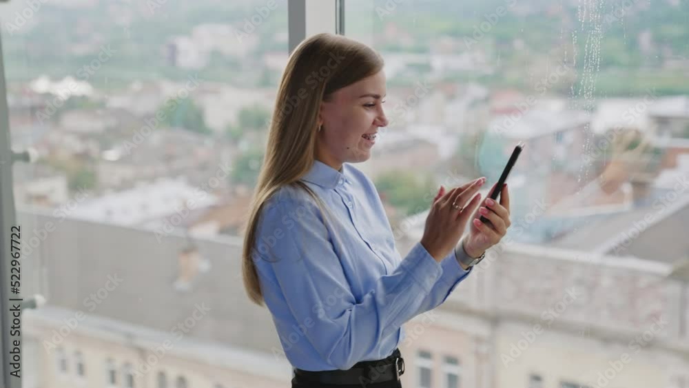 Happy smiling lady having video call on her smartphone. Woman talks on the phone standing near the window. Happy smiling lady having video call on her smartphone. Woman talks on the phone standing near the window.