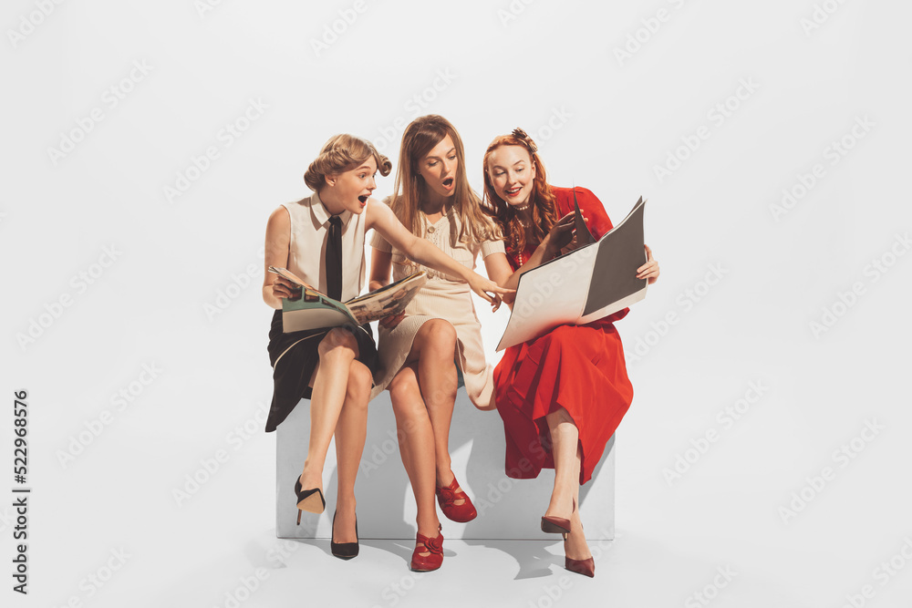 Naklejka premium Portrait of three young, beautiful, stylish woman sitting together and reading magazine isolated over white studio background