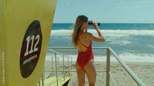 Young woman lifeguard on spanish beach look out through binoculars at sea. Bay watch concept, safety in open waters, safe vacation during storm. Sign says 112 emergency services in spanish photo