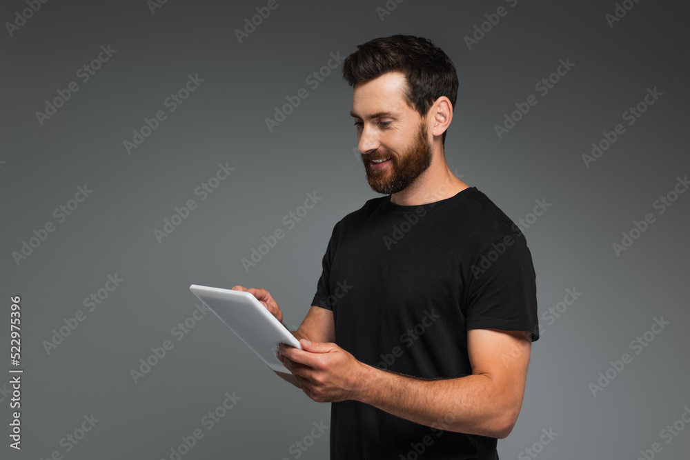 cheerful bearded man in black t-shirt using digital tablet isolated on grey. cheerful bearded man in black t-shirt using digital tablet isolated on grey.