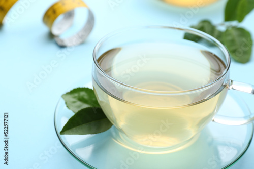 Glass cup of diet herbal tea and green leaves on light blue background, closeup