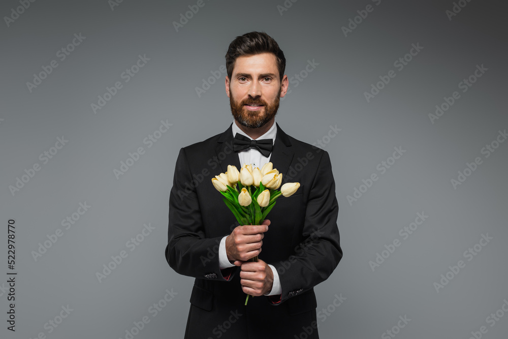 bearded man in elegant suit with bow tie holding bouquet of tulips isolated on grey. bearded man in elegant suit with bow tie holding bouquet of tulips isolated on grey.