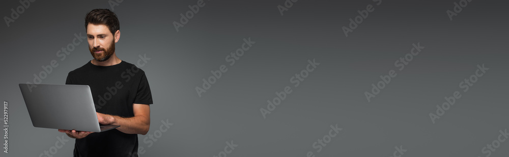bearded man in black t-shirt using laptop isolated on grey, banner. bearded man in black t-shirt using laptop isolated on grey, banner.