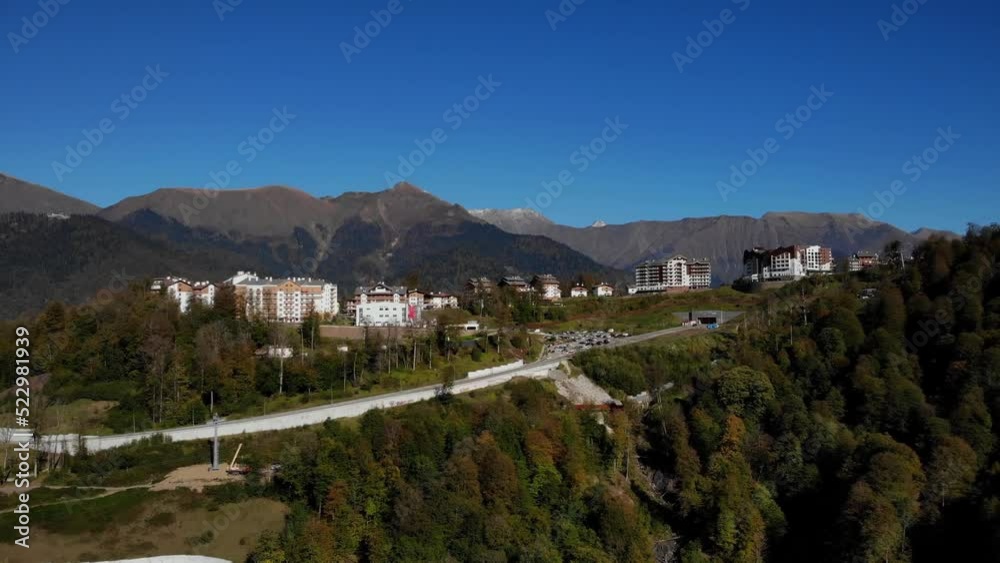 Autumn landscape of rosa khutor village among the caucasus mountains. sharp mountain peaks against blue sky. forest trees in colourful fall colors.