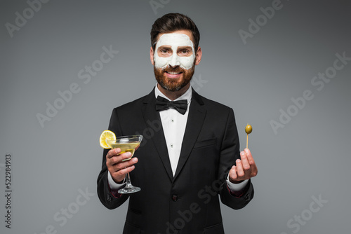 Obraz na płótnie happy man in suit with clay mask on face holding cocktail and toothpick with olive isolated on grey