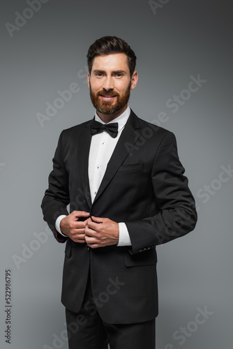 Fotografie bearded man in black tuxedo with bow tie smiling isolated on grey