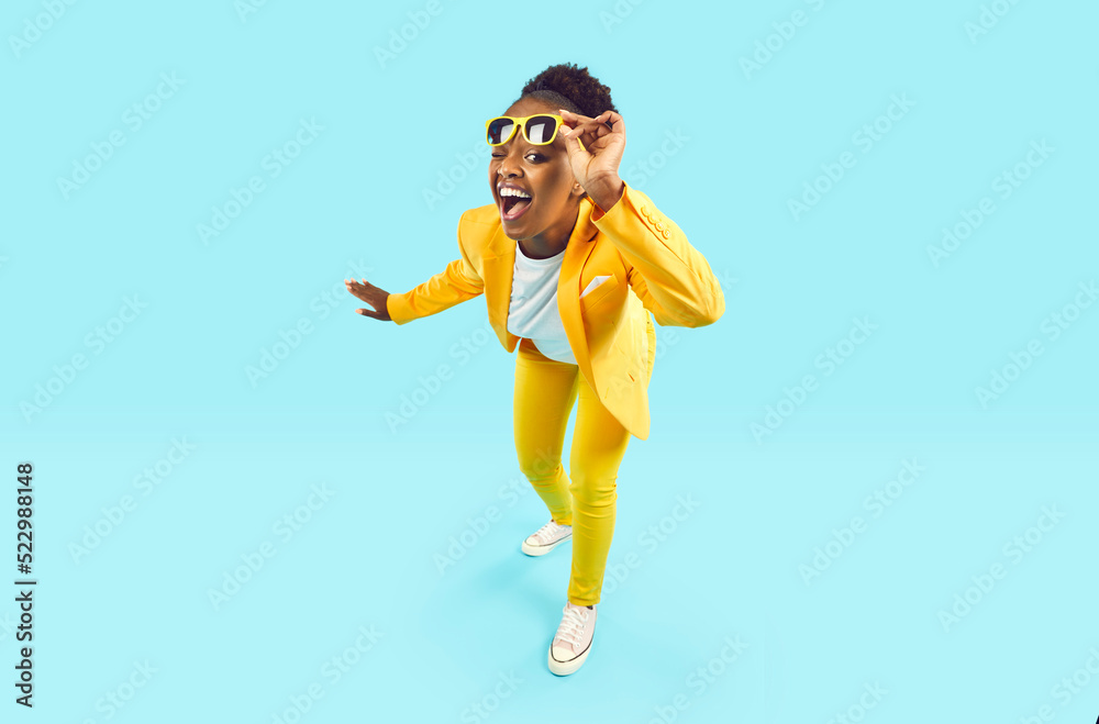 Obraz premium Full body happy cheerful Afro American woman wearing stylish yellow suit and trendy glasses standing isolated on blue background, looking at camera, winking her eye and smiling. Party, fashion concept