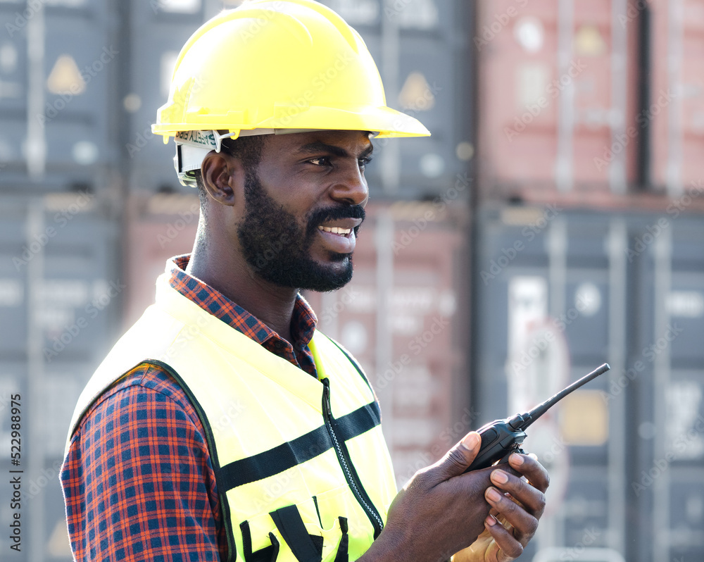 Industrial engineer man using walkie-talkie at container storage warehouse. African foreman wears safety helmet working at International logistic shipping port controls import export courier delivery