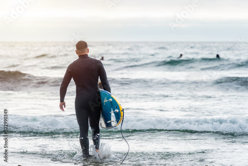 Surfer entering the sea carrying the surfboard looking at other surfers. High quality photo