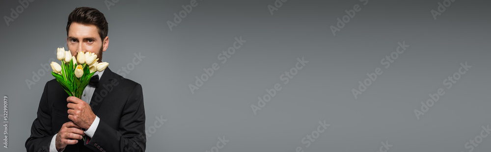 bearded man in elegant tuxedo smelling fresh tulips isolated on grey, banner. bearded man in elegant tuxedo smelling fresh tulips isolated on grey, banner.