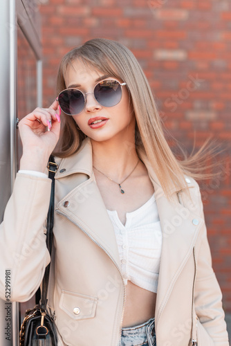 Fashionable young beautiful woman in casual fashion clothes with leather white jacket and bag wears a stylish round sunglasses, walks in the city near a brick wall