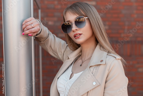Female fashion portrait of a beautiful young zoomer girl with round vintage sunglasses and a white leather jacket stands near a metal ads stand in the city © alones
