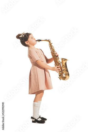 Charming little girl playing saxophone isolated on white studio background. Retro vintage style concept. Friendship, hobbies, art, eras comparison photo