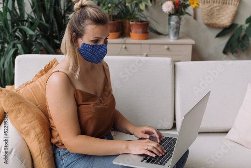 Closeup of young blonde woman with a protective face mask working on laptop in workshop, artist studio. Illness prevention.