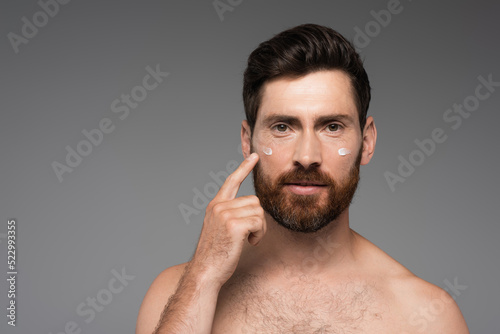 bearded man pointing with finger at cream on face isolated on grey.