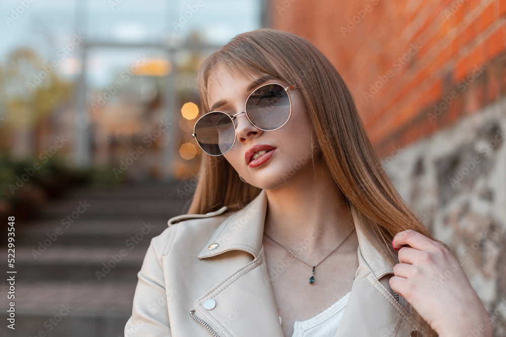 Urban female stylish portrait of a beautiful fashionable woman with vintage round hipster sunglasses in a leather jacket sits near a vintage mall on the street