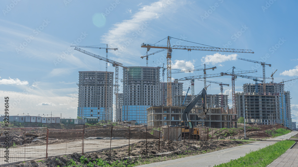 Construction of a multi-story residential buildings. Defocused foreground with young tree. Cranes work. Construction site. Construction of a multi-story residential buildings. Defocused foreground with young tree. Cranes work. Construction site.