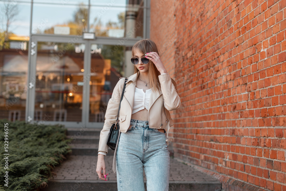 Fashion stylish lifestyle urban woman in trendy leather jacket and vintage jeans wears a fashionable sunglasses and walks in the city