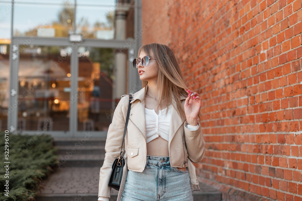 Street beautiful fashion woman with round sunglasses in leather jacket and bag with jeans walks in the city