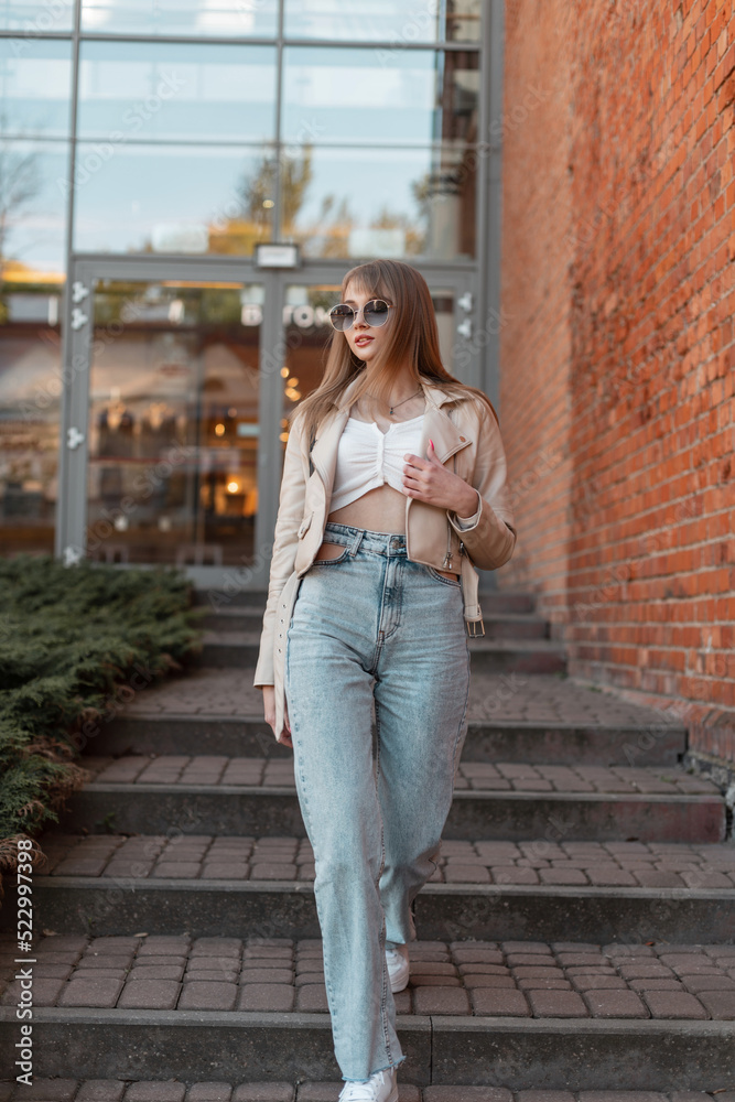 Stylish beautiful young woman with trendy sunglasses in a fashionable beige leather jacket with fashion vintage high waist jeans and white sneakers walks near the brick shopping mall