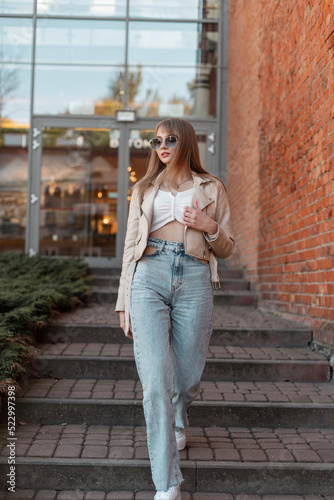 Stylish beautiful young woman with trendy sunglasses in a fashionable beige leather jacket with fashion vintage high waist jeans and white sneakers walks near the brick shopping mall