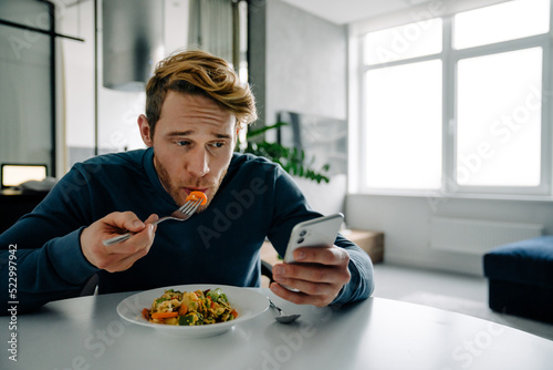 Young bearded white man eating dinner and using cellphone at home