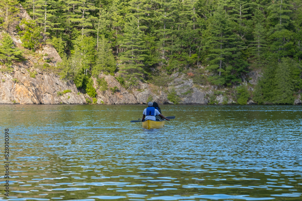 Couple in life vests paddling in a bright yellow kayak on a lake surrounded by rocks and thick coniferous forest, sunny summer day, selective focus. Water sports, recreation, adventure concept.