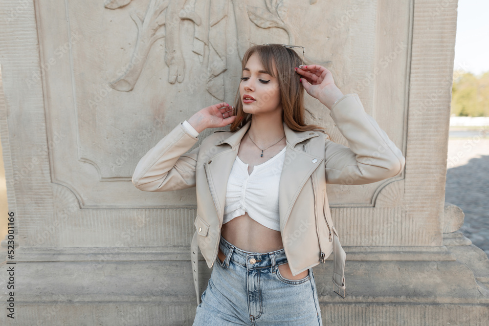 Stylish beautiful fashion young girl with a cute fresh face in a fashionable beige leather jacket with a top and jeans near a vintage column in the park. Female style outwear and beauty