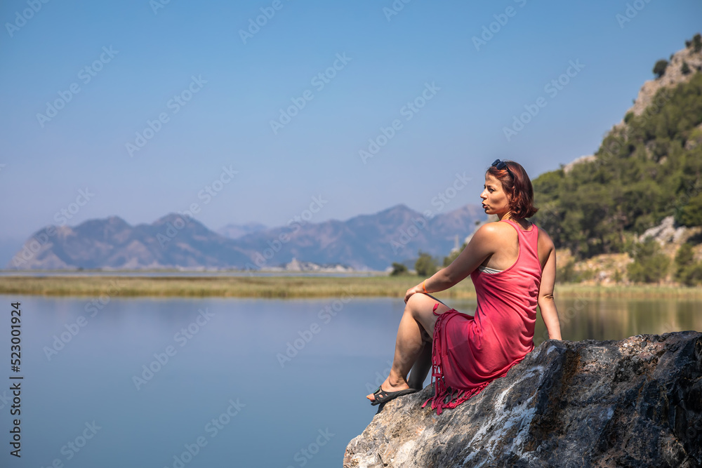 Beautiful girl in a red dress by the sea. Sunny day. Colorful sea landscape. Beautiful mountains. Girl by the sea. A girl in a dress. Summer day.