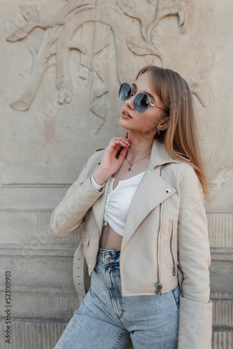 Fashion rock stylish beauty woman hipster with trendy sunglasses in fashionable leather jacket, jeans and white top poses near a vintage wall outdoors