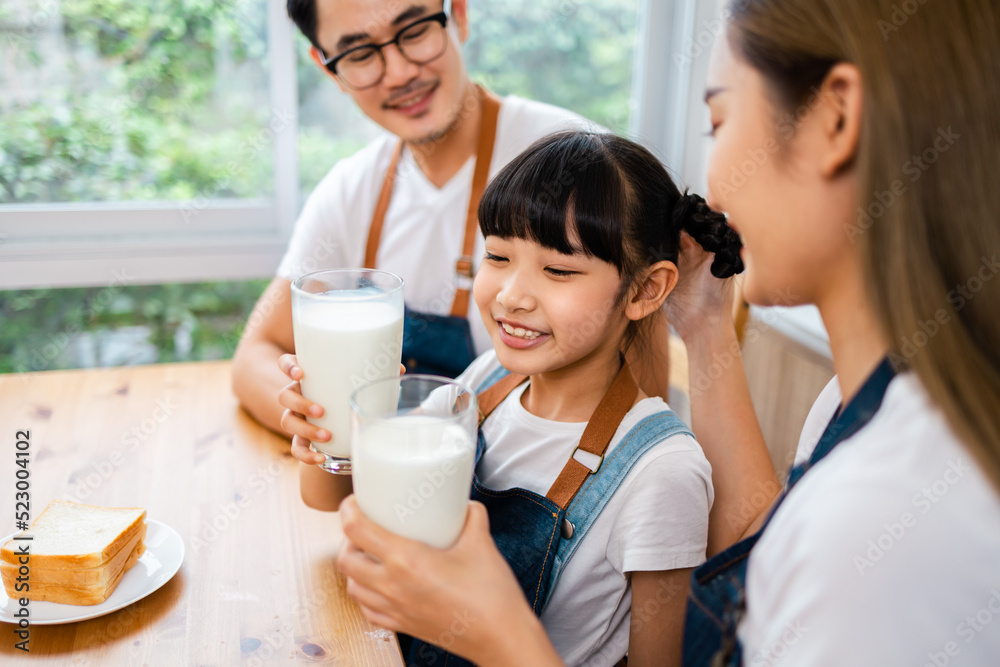 Asian family enjoying breakfast at cozy kitchen, little girl daughter sitting on table, drinking milk with smiling father and mother in morning. Happy family in kitchen. Asian family enjoying breakfast at cozy kitchen, little girl daughter sitting on table, drinking milk with smiling father and mother in morning. Happy family in kitchen.