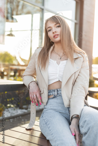 Fashionable beautiful woman in casual rock style clothes with leather jacket, top and jeans sits on wooden bench in the city at sunset