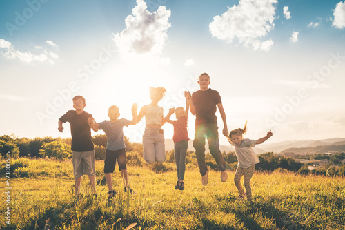 Fotografie Six kids brothers and sisters teenagers and little kids jumping on a green grass meadow with evening sunset background light holding hands in hands and smiling