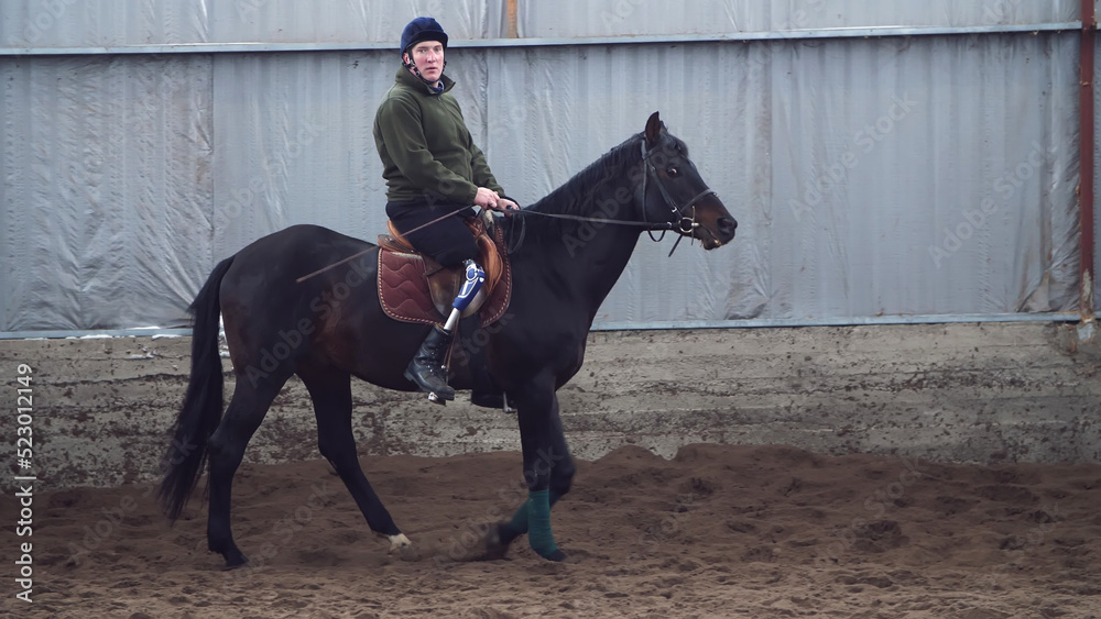 in special hangar, a young disabled man learns to ride a black, thoroughbred horse, hippotherapy. man has an artificial limb instead of his right leg. concept of rehabilitation of disabled with in special hangar, a young disabled man learns to ride a black, thoroughbred horse, hippotherapy. man has an artificial limb instead of his right leg. concept of rehabilitation of disabled with
