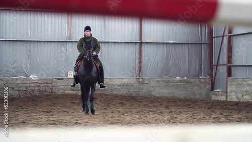 in special hangar, a young disabled man learns to ride a black, thoroughbred horse, hippotherapy. man has an artificial limb instead of his right leg. concept of rehabilitation of disabled with