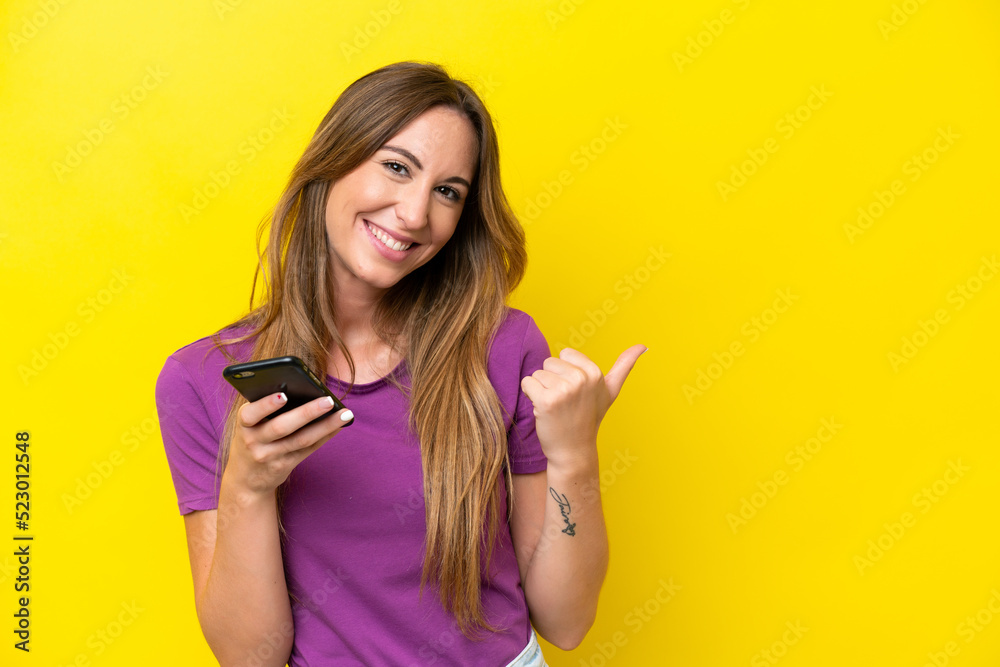Young caucasian woman isolated on yellow background using mobile phone and pointing to the lateral Young caucasian woman isolated on yellow background using mobile phone and pointing to the lateral