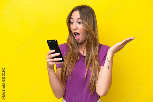 Young caucasian woman isolated on yellow background looking at the camera while using the mobile with surprised expression