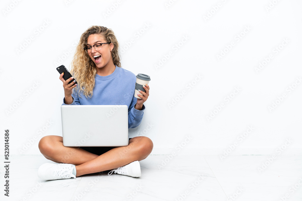 Girl with curly hair with a laptop sitting on the floor holding coffee to take away and a mobile